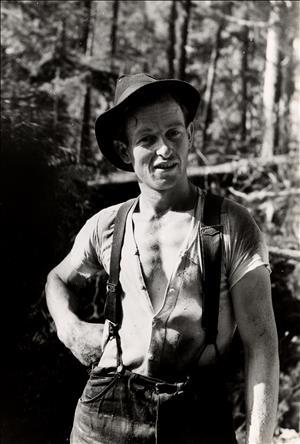 A young man stands with his hand on his hip. His shirt is unbuttoned showing his chest. He wears a floppy hat and suspenders. He is in the forest. 